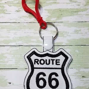 Pode incluir: Um chaveiro de plástico vermelho com uma placa branca da Rota 66 com letras e borda pretas.