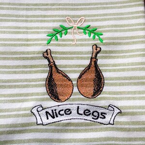 May include: A white and green striped kitchen towel with embroidered images of two roasted chicken legs and a banner that reads "Nice Legs". A green sprig and bow are embroidered above the chicken legs.