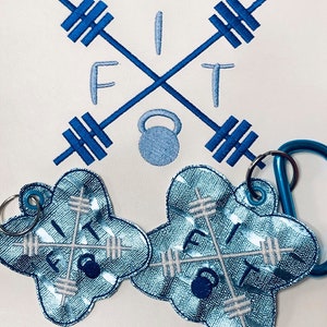 Może przedstawiać: Dwa niebiesko-białe haftowane breloki z motywem hantli i kettlebell. Breloki mają kształt chmury i napis "FIT" na nich.