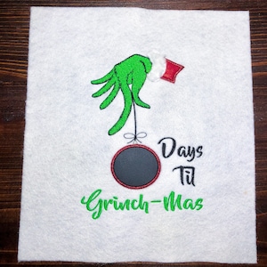 May include: Green Grinch hand holding a red and black Christmas ornament with a blank space for writing. The text "Days Til Grinch-Mas" is embroidered below the ornament.