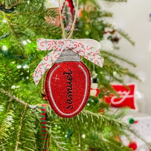 May include: A red Christmas light bulb ornament with the name "Samuel" embroidered in black script. The ornament is trimmed with silver thread and has a white ribbon with red and green candy cane stripes. It hangs from a natural twine string.