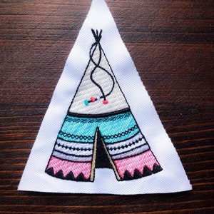 May include: A white fabric patch with an embroidered design of a teepee tent. The teepee is decorated with black, white, pink, and turquoise embroidery. The teepee has a black flap door and a black rope at the top.
