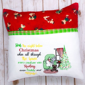 May include: White pillow with red and green plaid trim. The pillow has an embroidered design of a cat reading a book in a green armchair, a teddy bear, and a Christmas tree. The text reads "Twas the night before Christmas when all through the house every creature was Reading. even Teddy and"