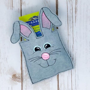 Könnte beinhalten: Grauer Filz-Osterkorb in Hasenform mit rosa Ohren und rosa Nase. Der Korb hat gestickte Augen und Schnurrhaare. Der Korb ist mit einer kleinen blauen und gelben Schachtel Schokolade gefüllt.