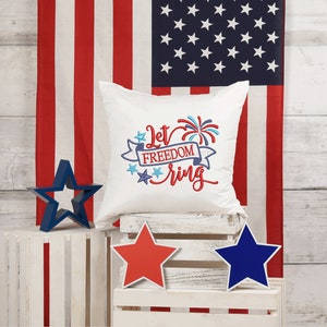 May include: White pillow with red, white, and blue embroidered text that says "Let Freedom Ring" with a firework design. The pillow is sitting on a white wooden crate with a red, white, and blue star on each side.