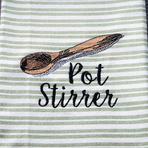 May include: A white and green striped kitchen towel with an embroidered wooden spoon and the words "Pot Stirrer" in black script. The towel is folded and placed on a dark wooden surface.