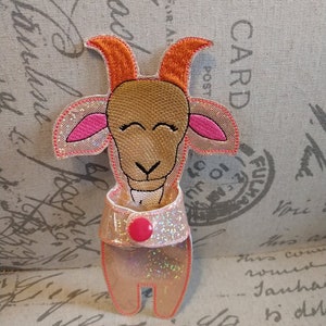 May include: A whimsical goat-shaped applique with a smiling face, orange horns, and pink ears. The goat has a sparkly, light pink body and a matching sparkly collar with a pink button. The applique is likely for crafts or embellishment.