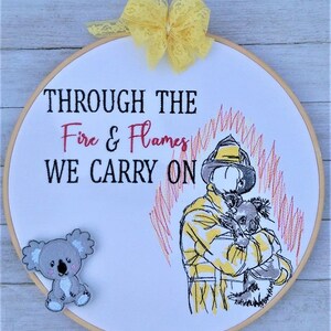 Può includere: Arte ricamata su un cerchio che rappresenta un pompiere che tiene un koala, con il testo "Through the Fire & Flames We Carry On". Un piccolo peluche di koala è attaccato al cerchio.