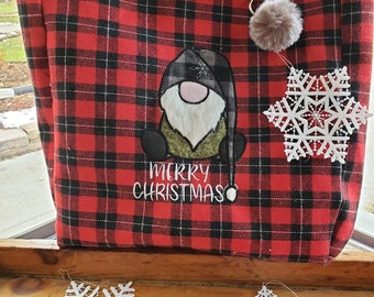 May include: Red and black plaid tote bag with a gnome applique and the text "MERRY CHRISTMAS". The bag has brown leather handles, a brown pom-pom, and a white snowflake ornament. The bag is sitting on a wooden surface.
