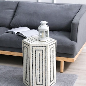 Könnte beinhalten: Ein Beistelltisch mit Mosaikmuster in Weiß und Grau mit geometrischem Design. Oben steht eine weiße Laterne mit Sternaussparungen. Der Tisch steht neben einem grauen Sofa und einem Buch. Der Tisch ist etwa 46 cm hoch.
