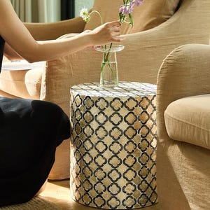 May include: A decorative side table with a geometric pattern in gold and black. The table has a cylindrical shape and is topped with a clear glass vase holding purple flowers. The table is next to a beige armchair.