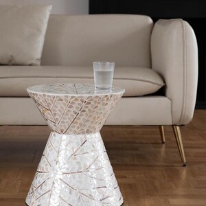 Könnte beinhalten: Ein dekorativer Beistelltisch mit einem Mosaik-Muscheldesign. Der Tisch hat ein weißes und beiges Farbschema mit einem Blattmuster. Ein Glas Wasser steht auf dem Tisch. Der Tisch steht neben einem beigen Sofa mit goldenen Beinen.