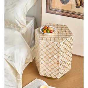 May include: A cream and gold mosaic patterned hexagonal side table. The table has a gold base and is topped with a small plate of tomatoes. The table is next to a bed with white bedding.
