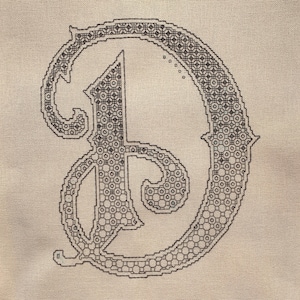 May include: A black and white embroidered letter D on a beige fabric background.