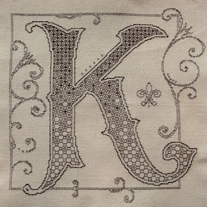 May include: A cross-stitch embroidery piece on light beige fabric featuring a large, ornate letter K in dark brown thread. The letter is filled with a geometric honeycomb pattern and surrounded by delicate, swirling filigree and a fleur-de-lis.