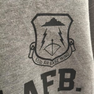 Pu&ograve; includere: Primo piano di una felpa grigia con una grafica nera. La grafica &egrave; uno scudo con una nuvola, fulmini e un arco e una freccia. &Egrave; visibile anche il testo "633d AIR BASE WING" e "AFB."