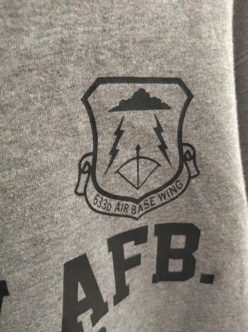 Pu&ograve; includere: Primo piano di una felpa grigia con una grafica nera. La grafica &egrave; uno scudo con una nuvola, fulmini e un arco e una freccia. &Egrave; visibile anche il testo "633d AIR BASE WING" e "AFB."