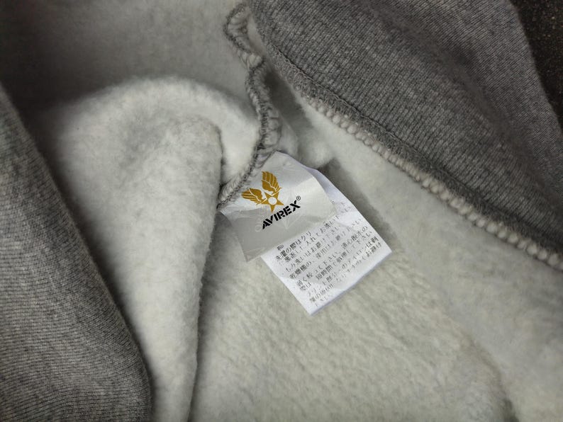 Pu&ograve; includere: Primo piano dell'interno di una felpa con cappuccio grigia con una morbida fodera bianca. La felpa con cappuccio presenta un'etichetta con il marchio "AVIREX" e un logo. Il tessuto sembra essere un materiale spesso e caldo, ideale per il freddo.