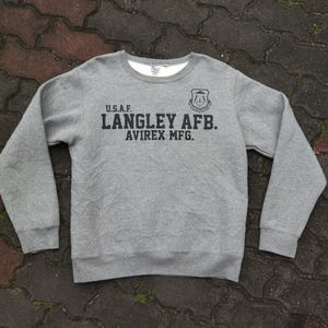 Pu&ograve; includere: Felpa grigia girocollo a maniche lunghe. Il davanti presenta la scritta "U.S.A.F. LANGLEY AFB. AVIREX MFG." in nero, insieme a un emblema nero. La felpa ha una fodera bianca intorno allo scollo.