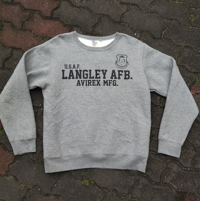 Pu&ograve; includere: Felpa grigia girocollo a maniche lunghe. Il davanti presenta la scritta "U.S.A.F. LANGLEY AFB. AVIREX MFG." in nero, insieme a un emblema nero. La felpa ha una fodera bianca intorno allo scollo.