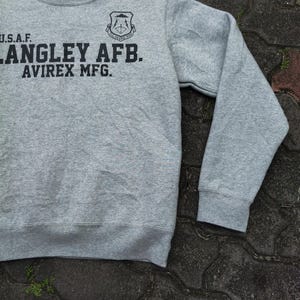 Pu&ograve; includere: Una felpa girocollo grigia con la scritta nera "U.S.A.F. LANGLEY AFB. AVIREX MFG.". Sopra il testo &egrave; presente un emblema nero. La felpa ha un colletto bianco e maniche lunghe.