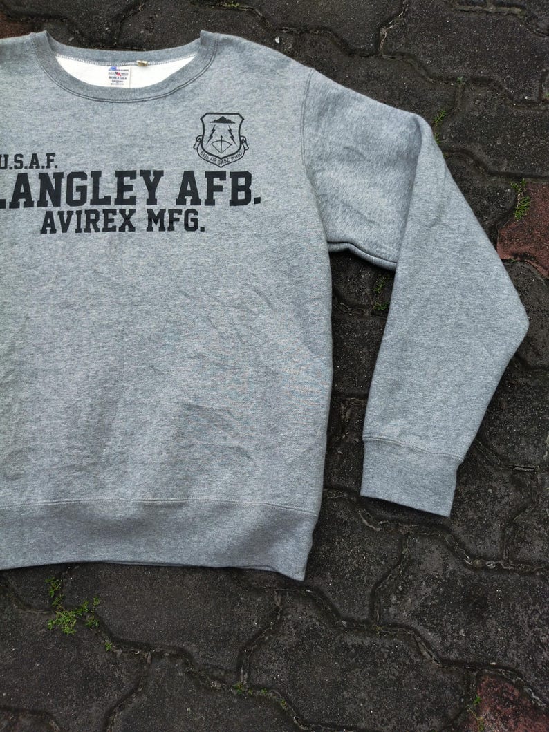 Pu&ograve; includere: Una felpa girocollo grigia con la scritta nera "U.S.A.F. LANGLEY AFB. AVIREX MFG.". Sopra il testo &egrave; presente un emblema nero. La felpa ha un colletto bianco e maniche lunghe.