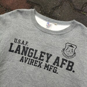 Pu&ograve; includere: Felpa girocollo grigia con testo nero "U.S.A.F. LANGLEY AFB. AVIREX MFG." e stemma nero. La felpa ha una fodera interna bianca intorno al colletto.