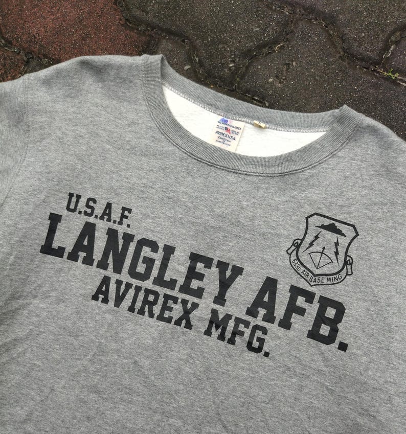 Pu&ograve; includere: Felpa girocollo grigia con testo nero "U.S.A.F. LANGLEY AFB. AVIREX MFG." e stemma nero. La felpa ha una fodera interna bianca intorno al colletto.