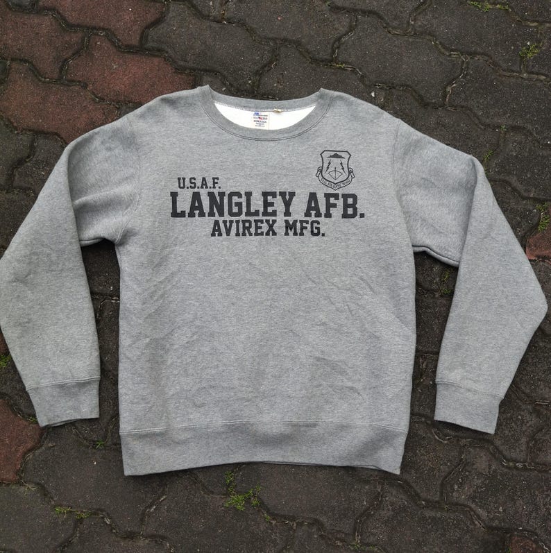 Pu&ograve; includere: Una felpa grigia girocollo con maniche lunghe. La parte anteriore presenta la scritta "U.S.A.F. LANGLEY AFB. AVIREX MFG." in nero, insieme a un emblema nero. La felpa ha una fodera bianca intorno allo scollo.