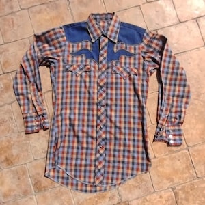 May include: A long-sleeved plaid shirt with a blue yoke and decorative stitching. The shirt has a button-down front, two chest pockets, and buttoned cuffs. The plaid pattern includes red, blue, and tan squares, creating a vintage Western style.