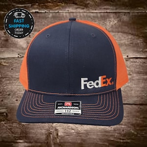 May include: A navy blue and orange trucker hat with a white "FedEx" logo embroidered on the front. The hat is adjustable and has a Richardson label.
