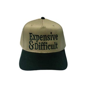 May include: A tan and black baseball cap with the embroidered text "Expensive & Difficult" in black. The cap has a black brim and a small black button on top. The hat is a flat-brim style.