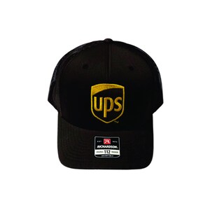 May include: A black trucker hat featuring a mesh back and a solid front panel with an embroidered yellow UPS shield logo. A rectangular sticker on the brim displays the text Richardson, Original 112 Trucker, Adjustable, Est 1970.