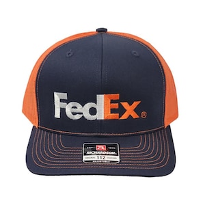 May include: A navy blue and orange trucker hat with the FedEx logo embroidered on the front. The hat features a mesh back, a curved brim, and an adjustable strap. The Richardson brand label is visible.