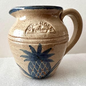 May include: A beige ceramic pitcher with a blue rim and handle. The pitcher has a blue pineapple design and the words "Old Shore Pottery" in a circle on the front.