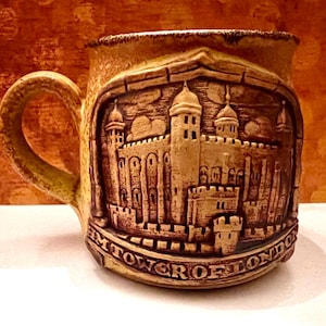 May include: A ceramic mug with a detailed relief of the Tower of London. The mug is tan with a darker brown relief and handle. The words "TOWER OF LONDON" are at the bottom of the relief.