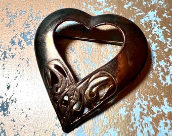 May include: A bronze-toned heart-shaped brooch with an open center and intricate filigree detailing on the lower left side. The brooch has a pin closure on the back. The metal has a slightly aged appearance.