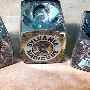 May include: Three vintage Sylvania Magicube flash cubes. The cubes have a clear plastic top with a silver reflector and a lower section with the word "Focal" printed on them. The center cube has a Sylvania Magicube label.