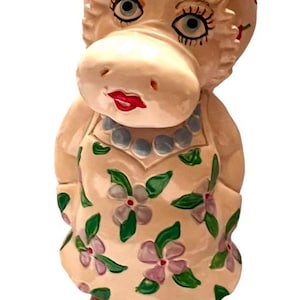 May include: A whimsical ceramic hippo figurine. The hippo wears a floral apron and a blue beaded necklace. The hippo's head is adorned with strawberries. The figurine has painted blue eyes and red lips. The hippo is standing on its feet.