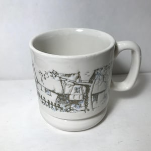 May include: White ceramic mug with a handle, featuring a black and blue illustration of a village scene. The mug has a rounded base and a cylindrical body. The handle is a simple, curved design.