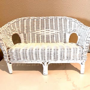 May include: A small, white wicker loveseat with a curved back and armrests. The seat has two openings on either side. The woven design is visible throughout the piece. The loveseat is elevated on four short legs.