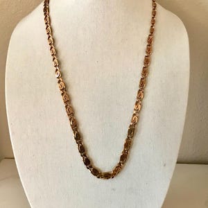 May include: A gold-toned necklace with a repeating, stylized S-link design. The necklace is displayed on a white mannequin bust. The chain is a warm, copper color, and the links are evenly spaced, creating a symmetrical pattern.