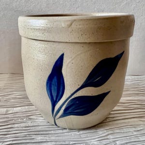 May include: A white ceramic crock with a blue leaf design. The crock has a wide mouth and a rounded body.