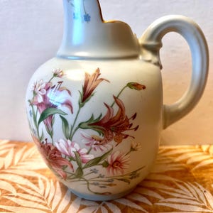 May include: A white ceramic pitcher with a floral design. The pitcher has a gold rim and a handle. The flowers are pink, orange, and green.