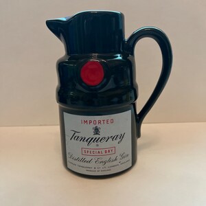 May include: Black ceramic pitcher shaped like a Tanqueray gin bottle. The pitcher has a red cap and the label reads "IMPORTED Tanqueray SPECIAL DRY Distilled English Gin CHARLES TANQUERAY & CO LTD LONDON ENGLAND PRODUCE OF ENGLAND"