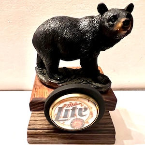May include: A black bear figurine standing on a dark wooden base. The bear has textured fur and a tan muzzle. Attached to the front of the wooden base is a circular Miller Lite beer logo with a gold rim and a black border, featuring the text Miller Lite.