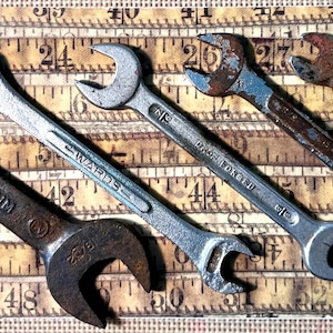 May include: Five vintage wrenches of varying sizes and wear, arranged diagonally. The wrenches are made of metal, with some showing signs of rust. The background is a vintage measuring tape with black numbers.
