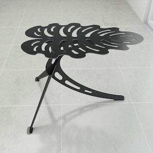 May include: Black metal side table with a leaf-shaped top. The table has a unique design with cut-out details and three legs. The table's dimensions are approximately 24 inches long, 18 inches wide, and 20 inches high.