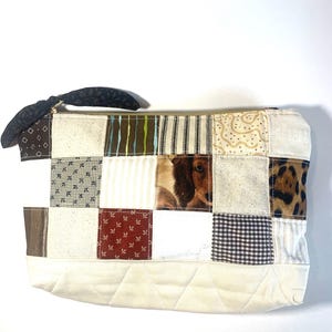May include: A patchwork zippered pouch featuring squares of various patterns and textures, including a dog portrait, stripes, and floral prints. The pouch has a black fabric loop and a quilted bottom.