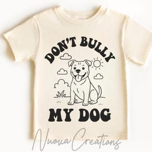 May include: Cream-colored t-shirt featuring the text "DON'T BULLY MY DOG" in black, with a smiling dog graphic. The design includes clouds, grass, and a sun. The brand name "Nuova Creations" is printed at the bottom.